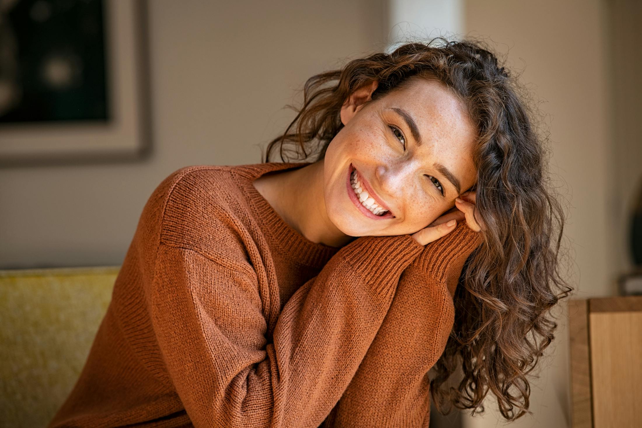 Woman with curly long brown hair in her 30's, smiles widely at the camera. Her hands are up by her cheek and she is wearing a cozy sweater