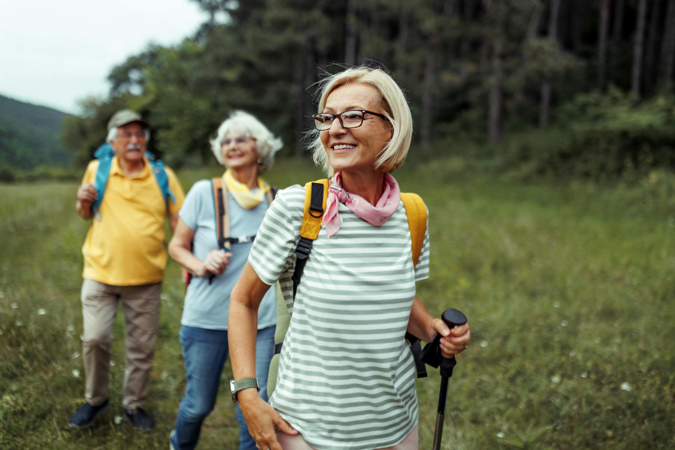 Two older women and one older man backpacking through a beautiful meadow lined with a forest full of trees. The hikers are all smiling enjoying the view.