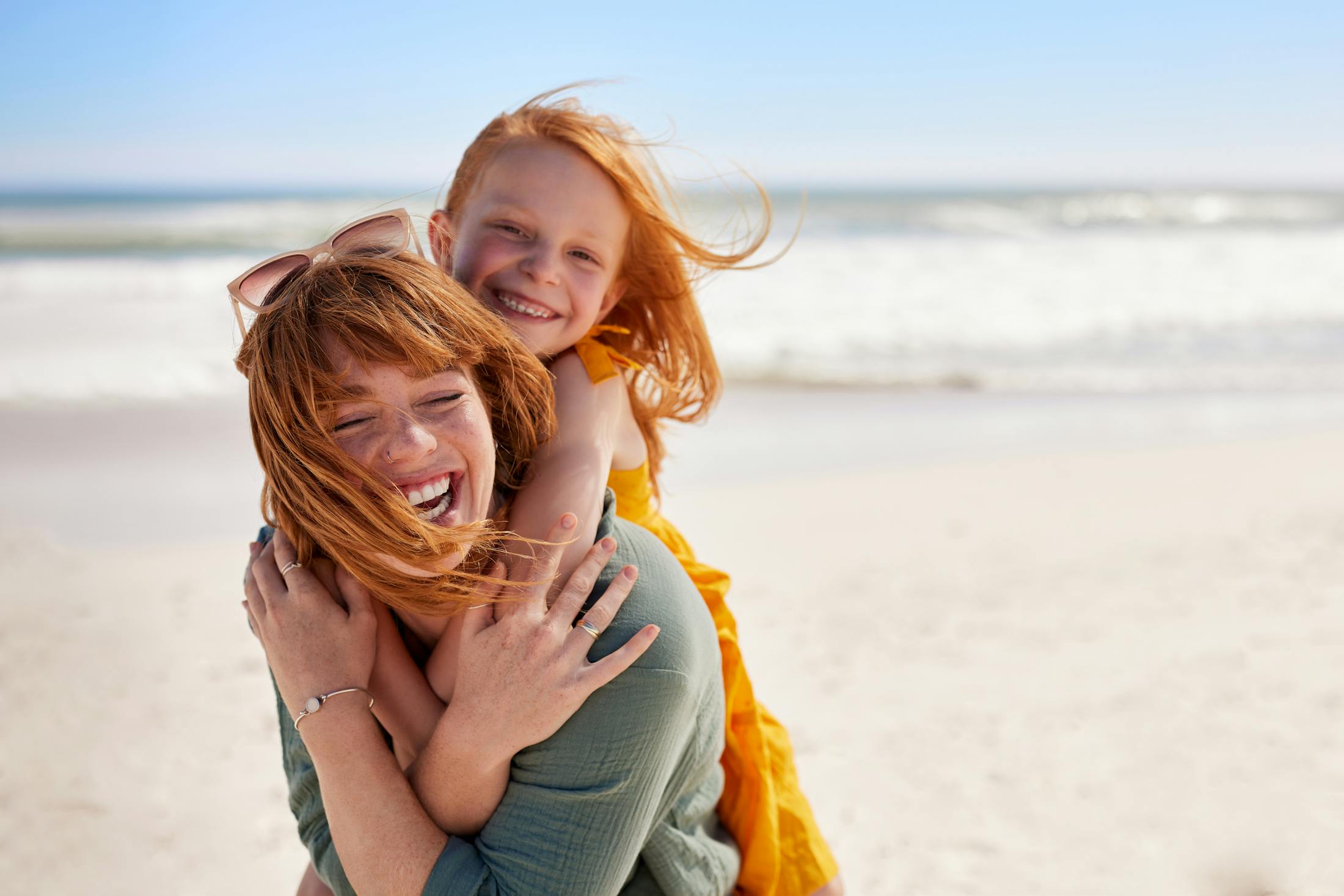 Woman smiling and laughing on a beach with her daughter on her back