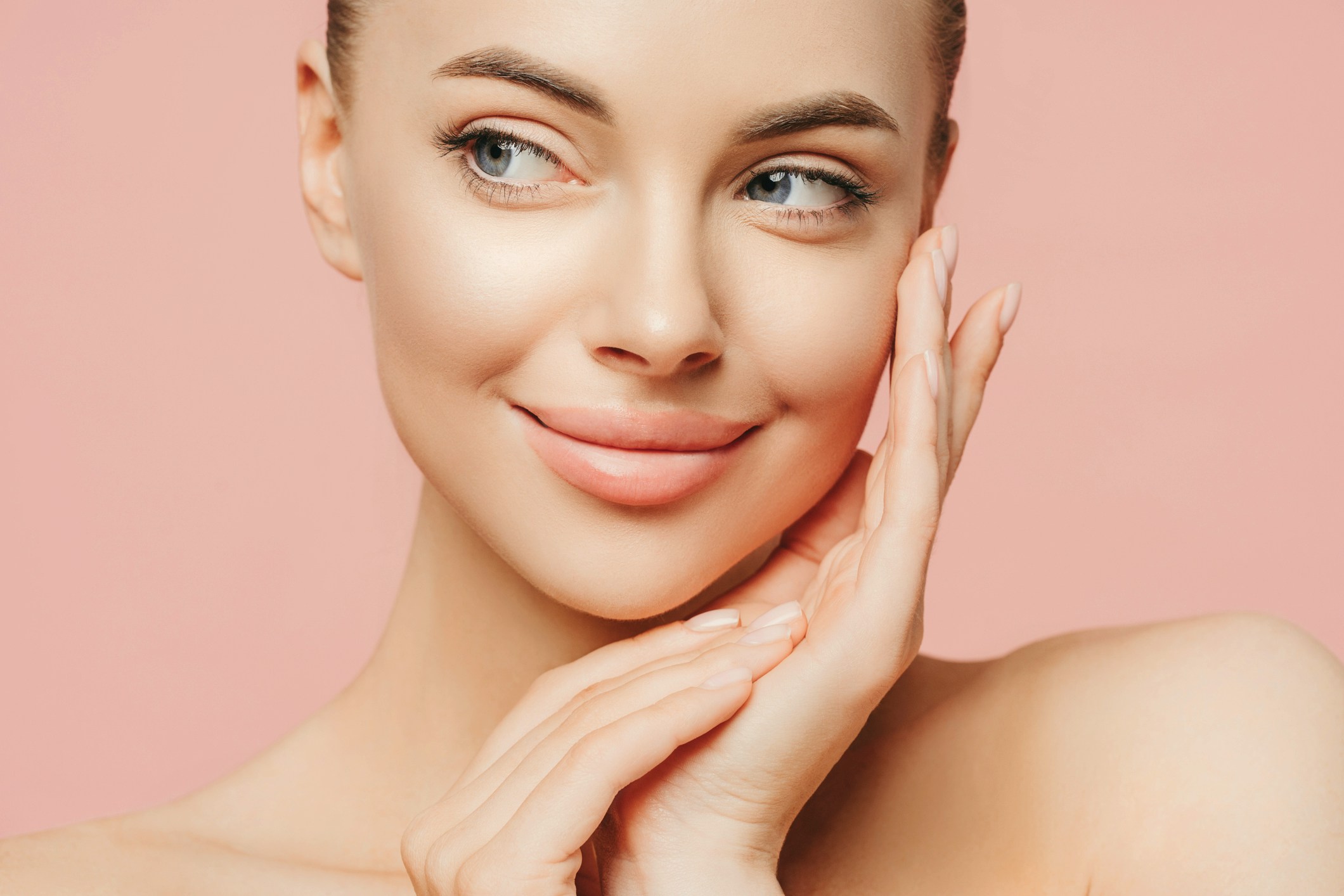 A woman with flawless skin holds her hands up to her cheek. She is in a studio with a pinnk background. You can only see her face and sholders. She is emulating what your skin may achieve with VAMP™ Salmon DNA Skin Rejuvenation