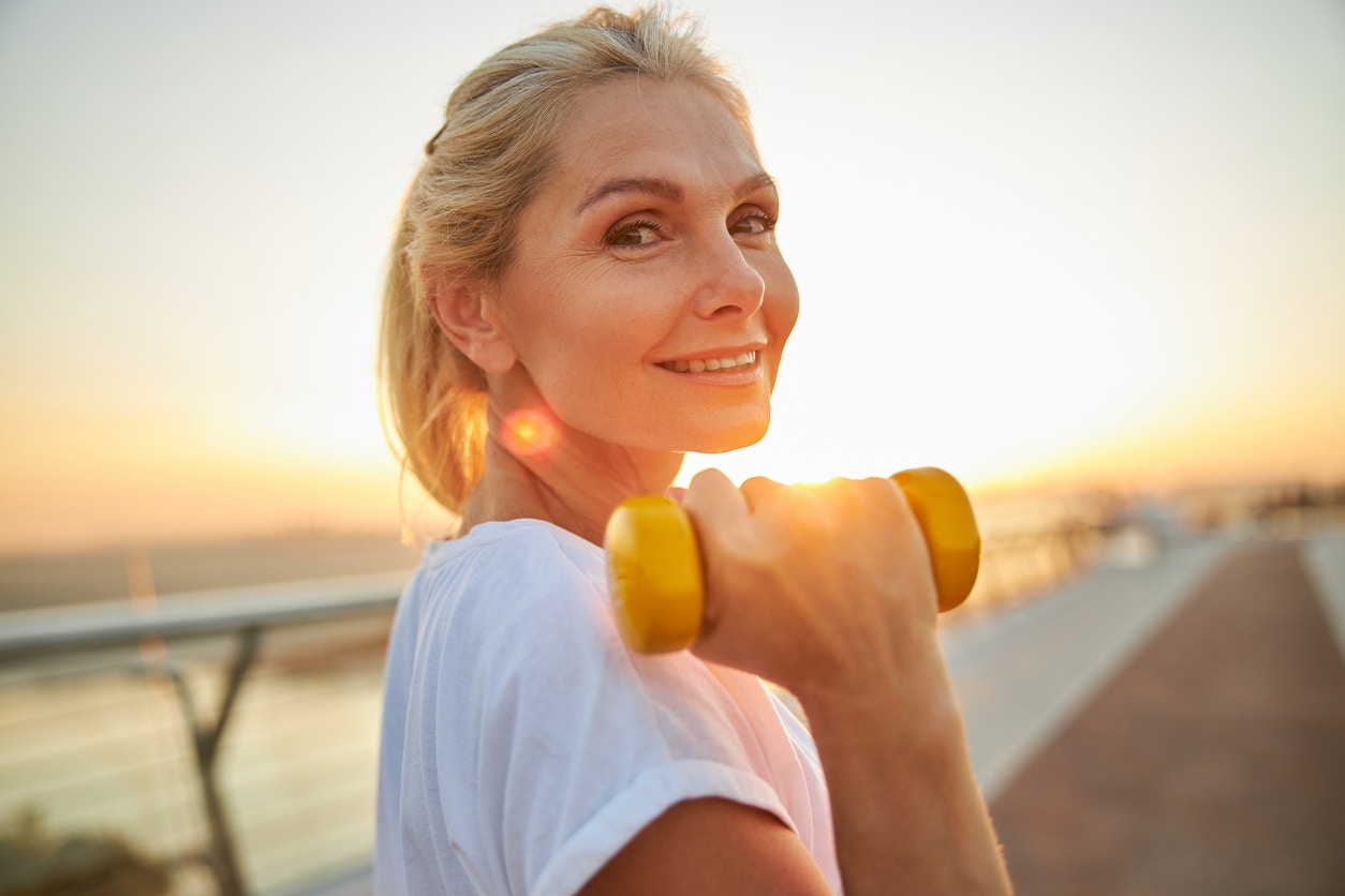 Older woman holding a weight in her hand outside while exercising, smiles at the camera. She had a youthful glow showing the potential results of a deep plane facelift in Beverly Hills with Dr. Torkian