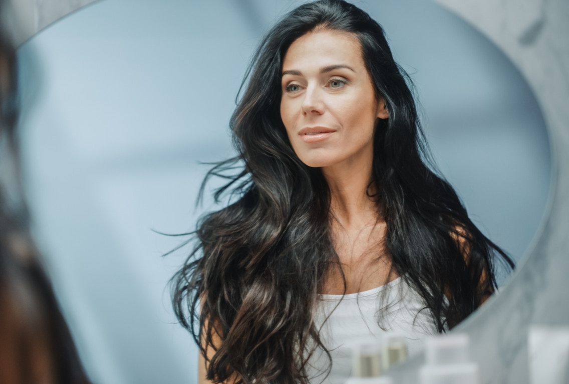 Woman in mid 40s with long hair looks at herself in the mirror with a small contented grin. Her youthful appearance, shows the potential results of a mini facelift in Beverly Hills with Dr. Torkian