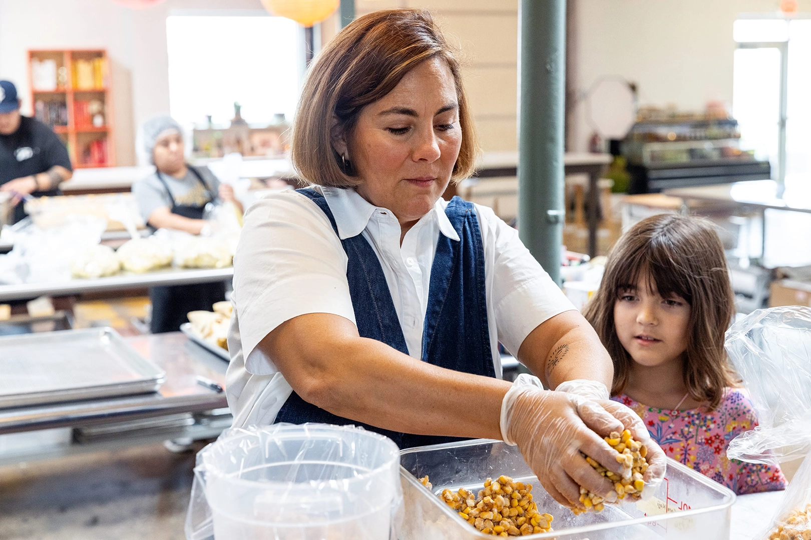 Woman moves handfuls of nixtamalized corn from one circular container into a rectangular container, while a little girl watches.