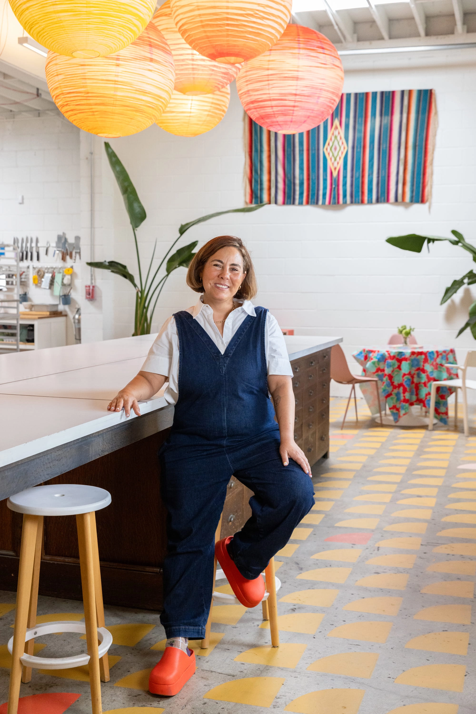 Smiling woman wearing jumpsuit sits on a stool inside a test kitchen.