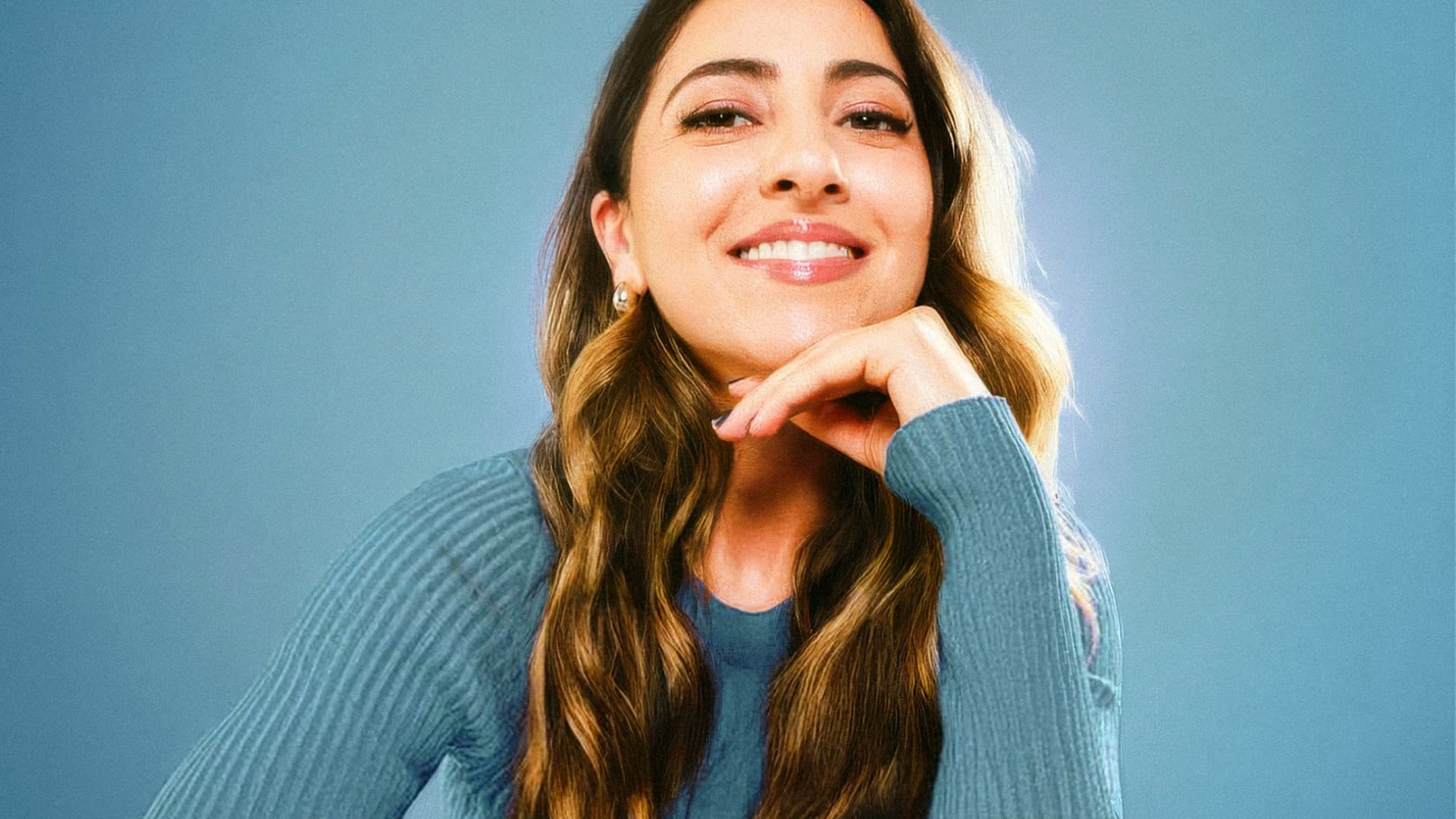 Smiling woman with long hair wearing blue sweater props her chin on her hand.
