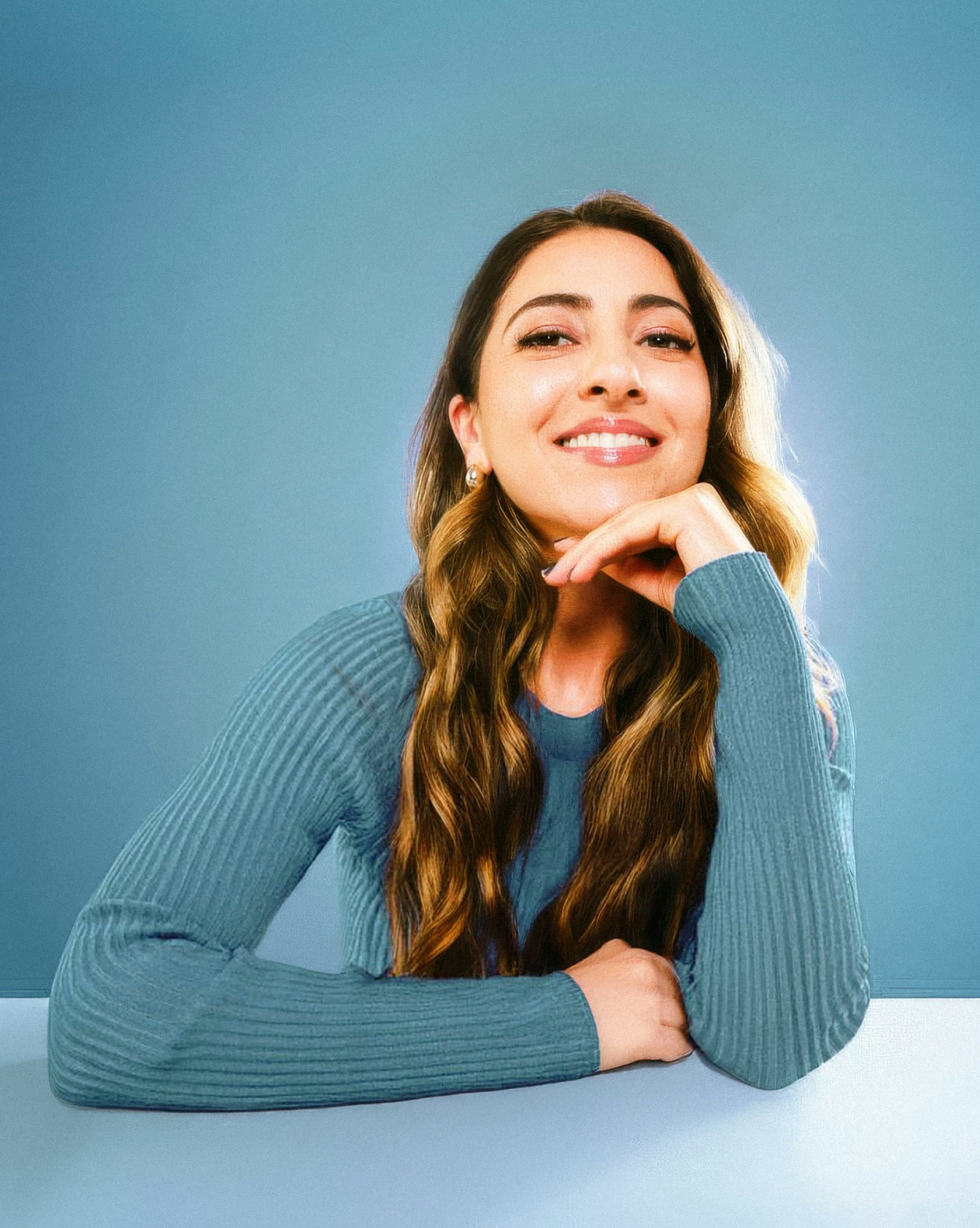 Smiling woman with long hair wearing blue sweater props her chin on her hand.