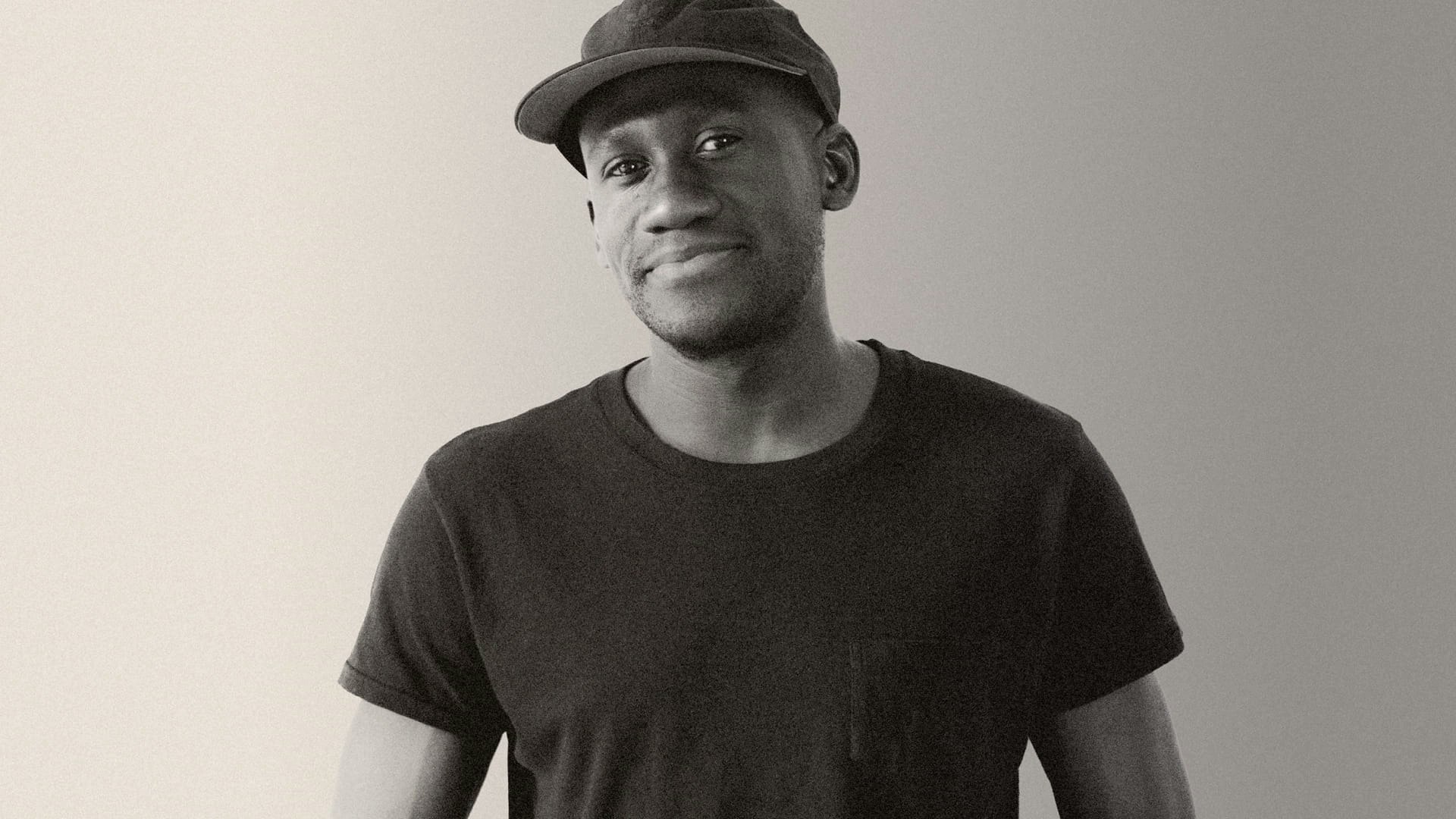 Smiling man wearing black cap and black t-shirt looks directly at the camera, with his hands in his pockets.