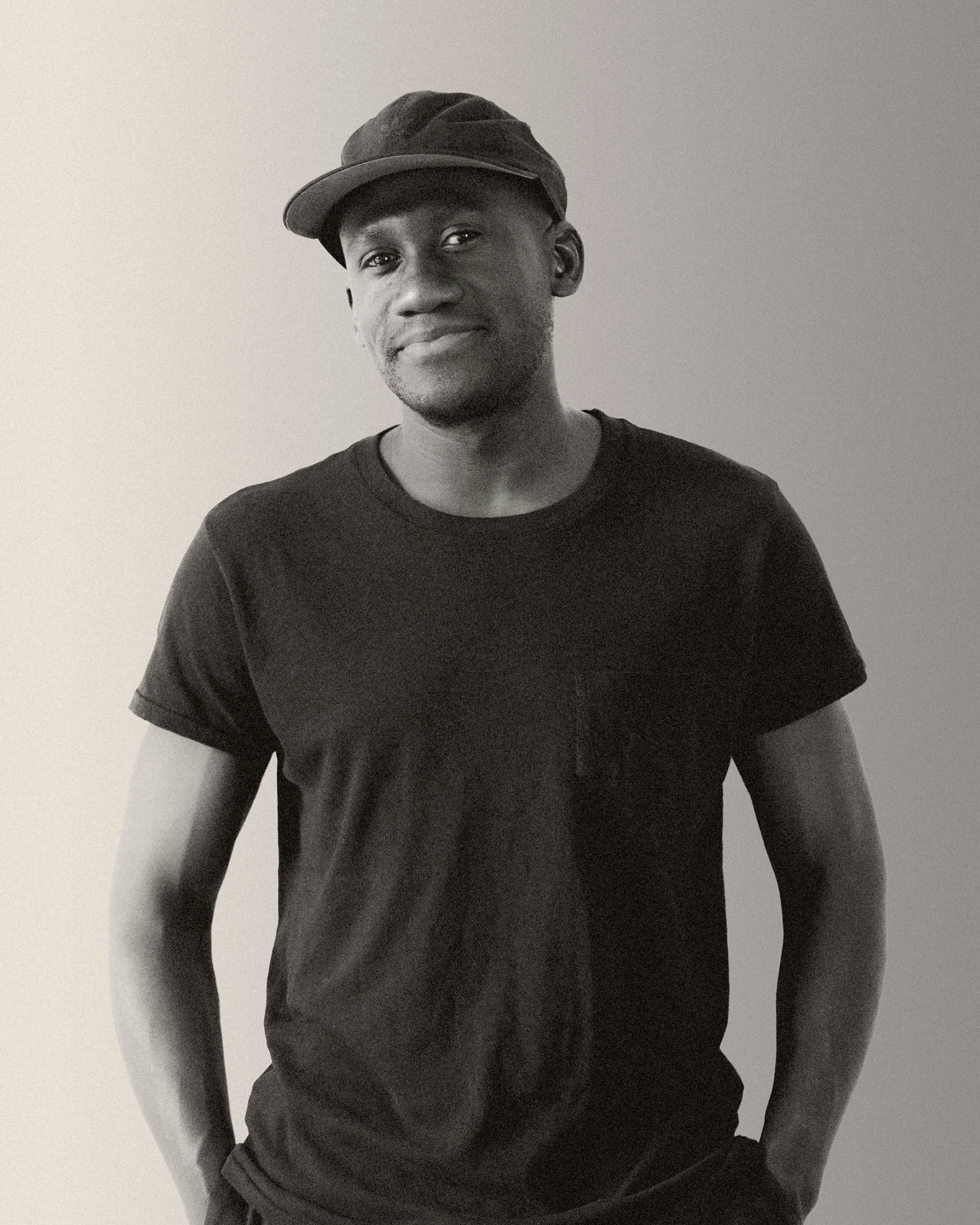 Smiling man wearing black cap and black t-shirt looks directly at the camera, with his hands in his pockets.