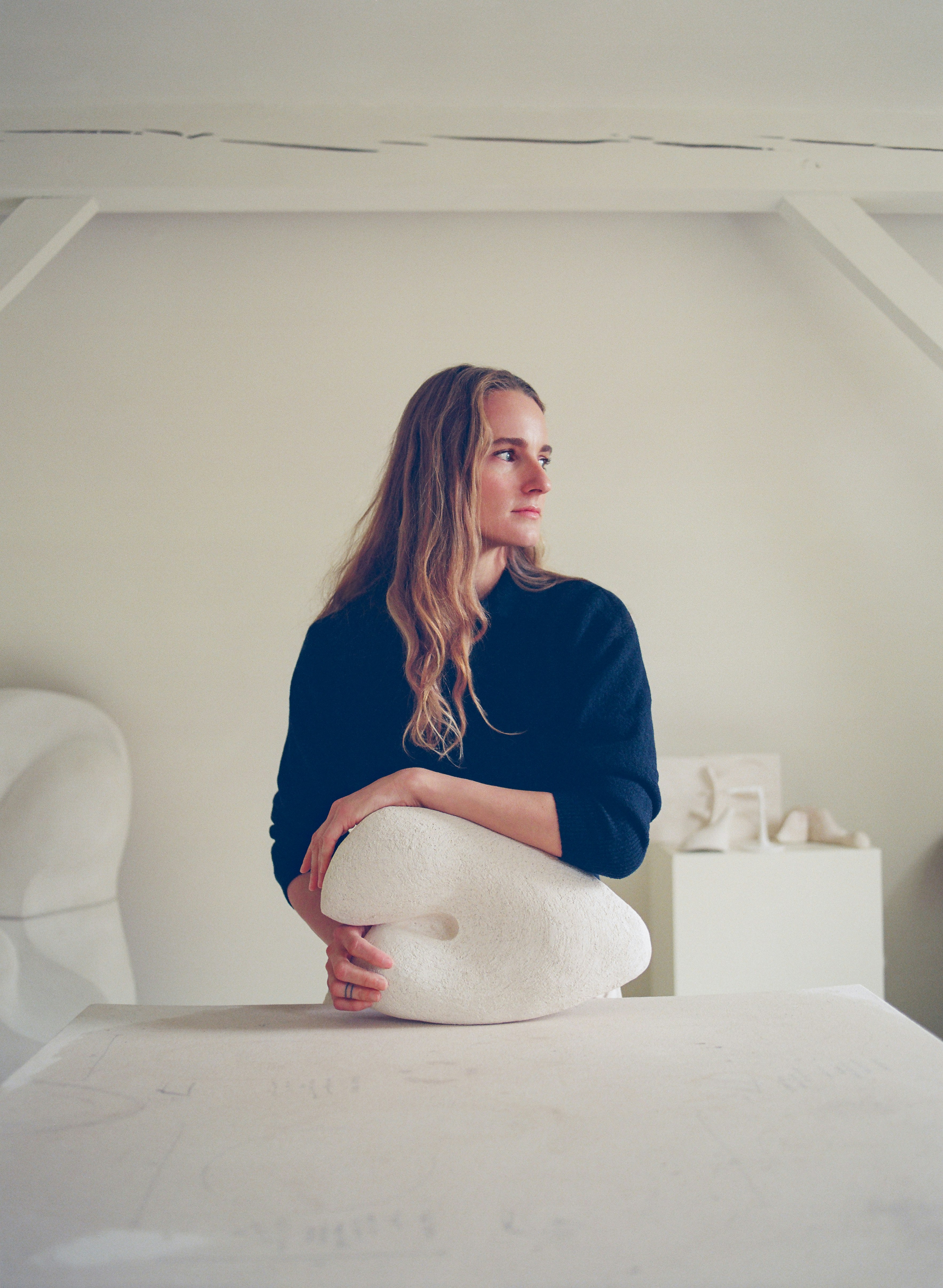 Woman inside an all-white studio space looks to her left while cradling a white sculpture in her hands.