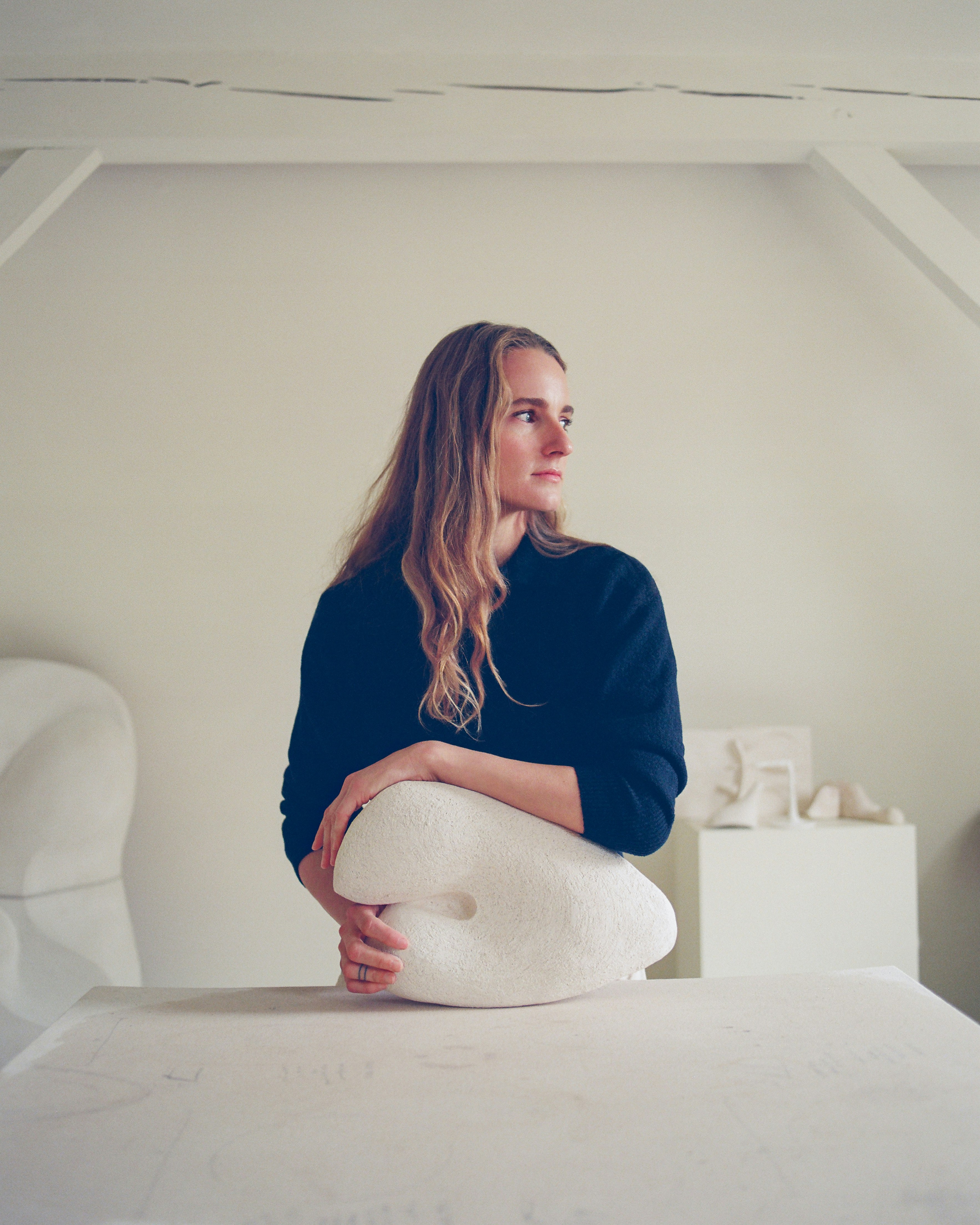 Woman inside an all-white studio space looks to her left while cradling a white sculpture in her hands.