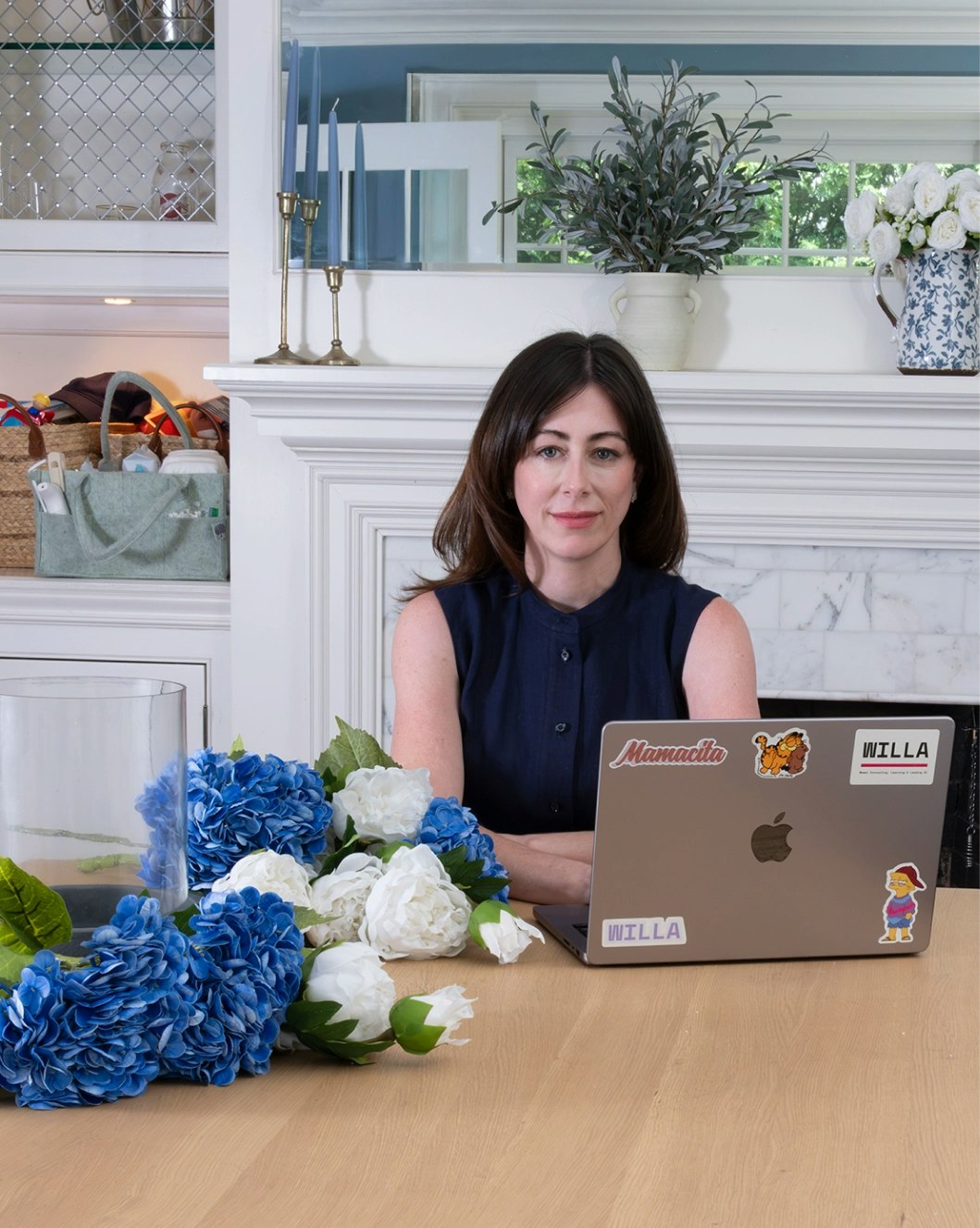 Woman looking directly at the camera sits at a table behind a laptop; next to her are cut blue and white flowers and an empty vase.