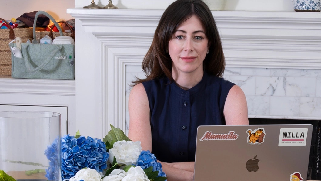 Woman looking directly at the camera sits at a table behind a laptop; next to her are cut blue and white flowers and an empty vase.
