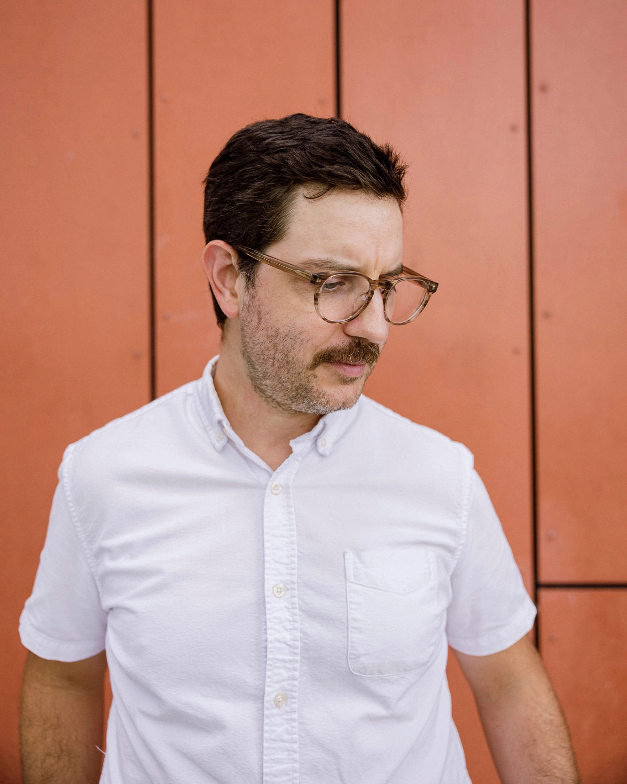Man with moustache wearing glasses looks slightly downward.
