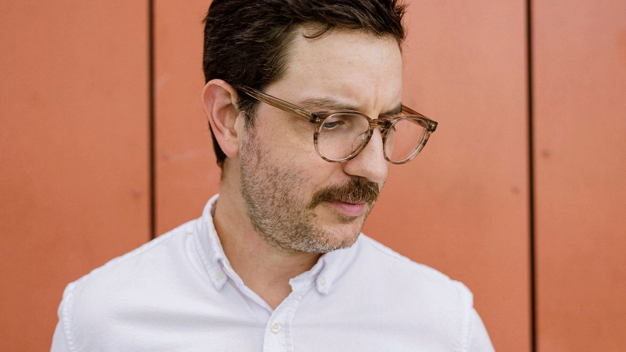 Man with moustache wearing glasses looks slightly downward.