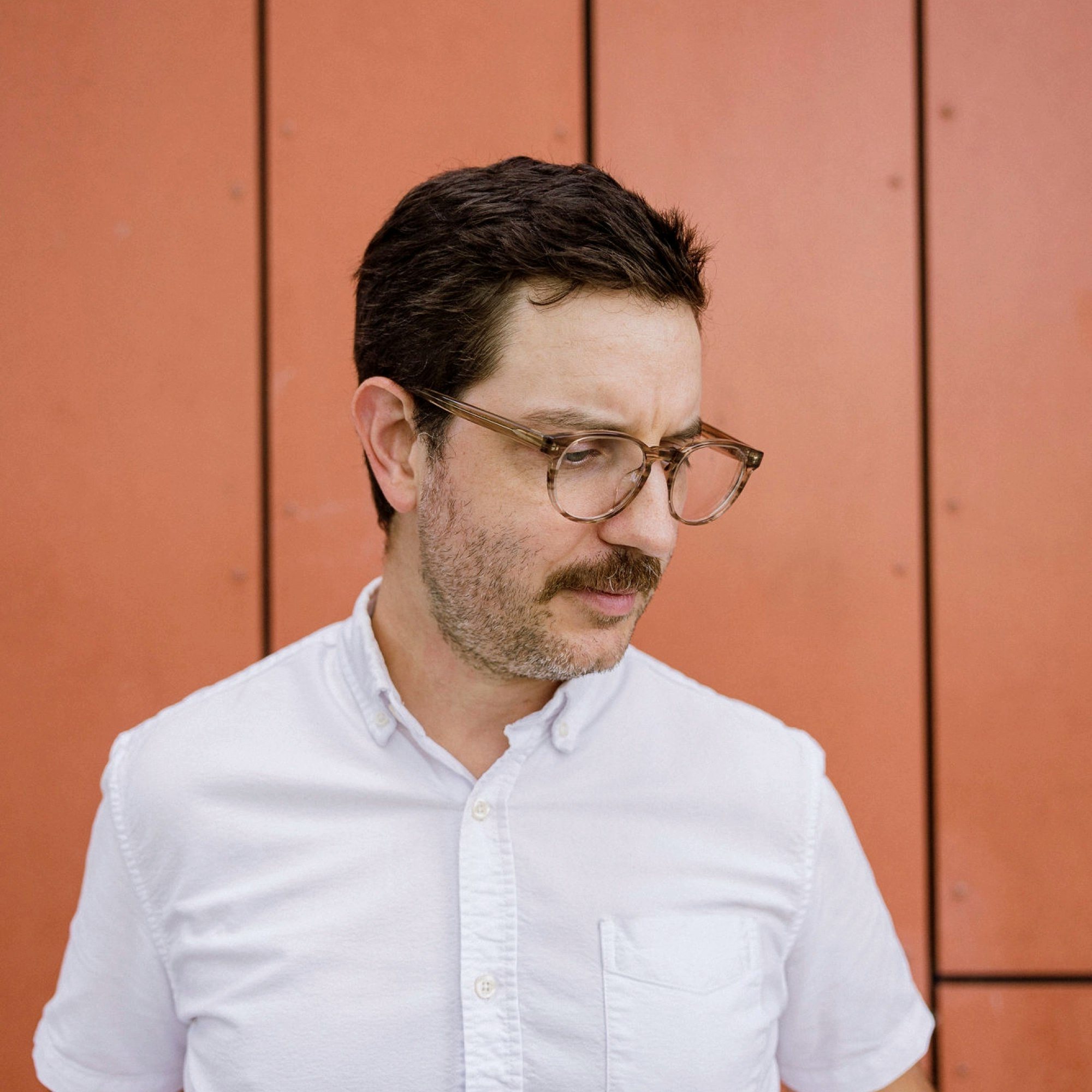 Man with moustache wearing glasses looks slightly downward.