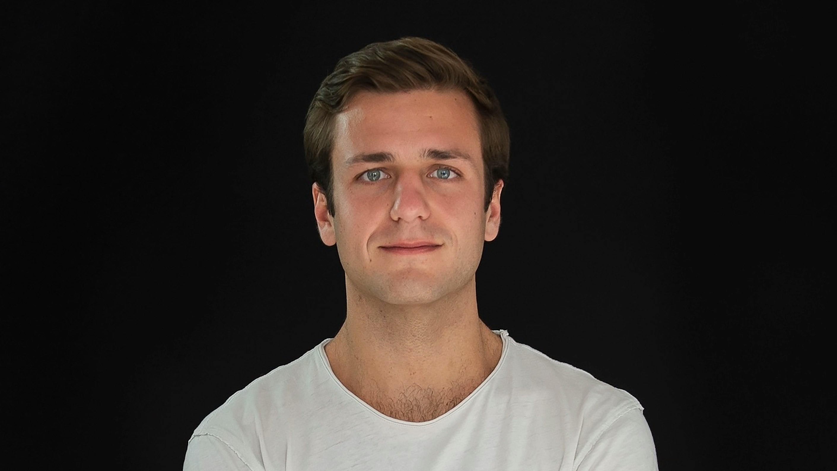 Man wearing white T-shirt and crossing arms looks directly at the viewer.