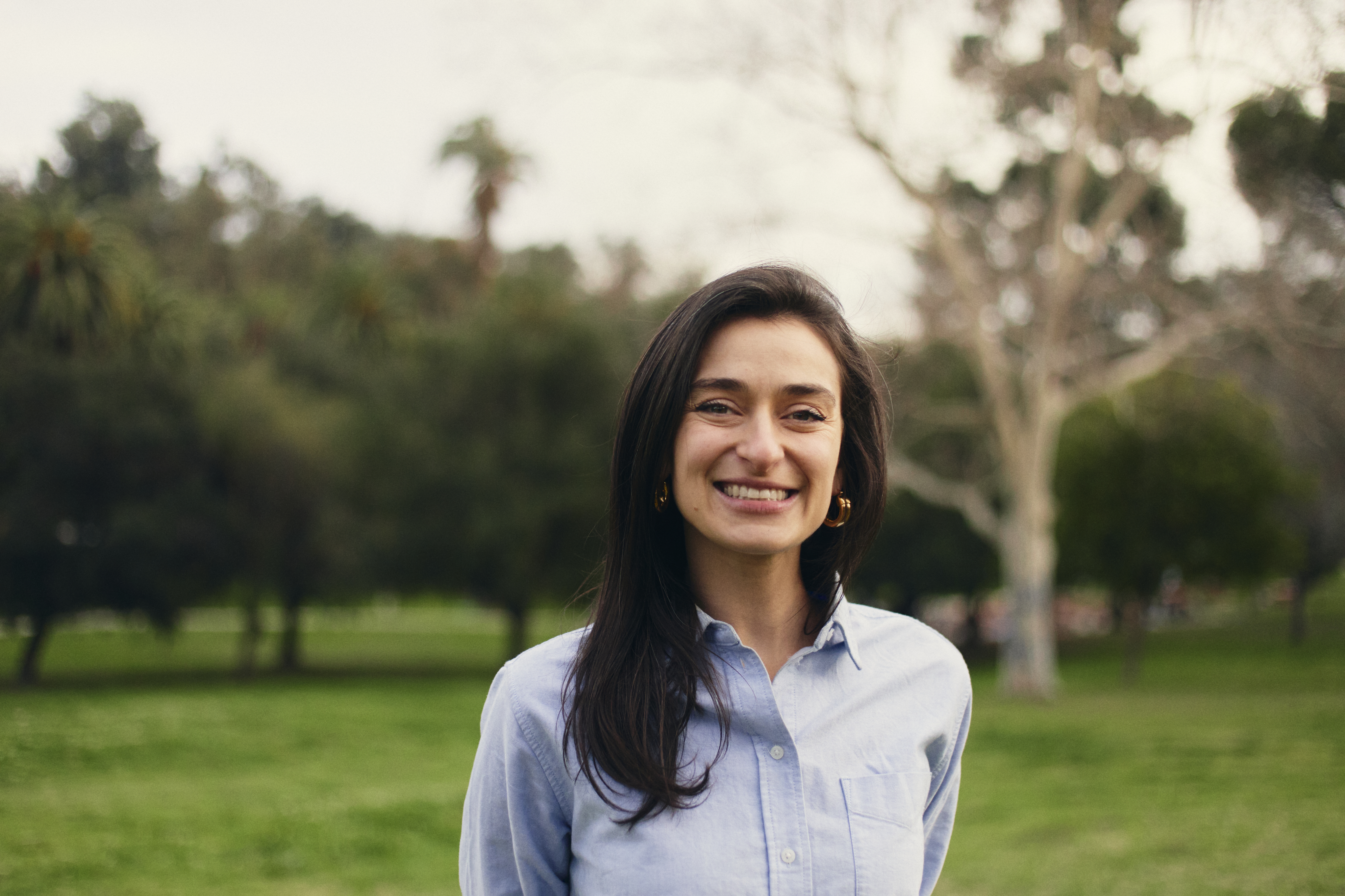 Smiling dark-haired woman wearing light blue button-down shirt stands in a green field.
