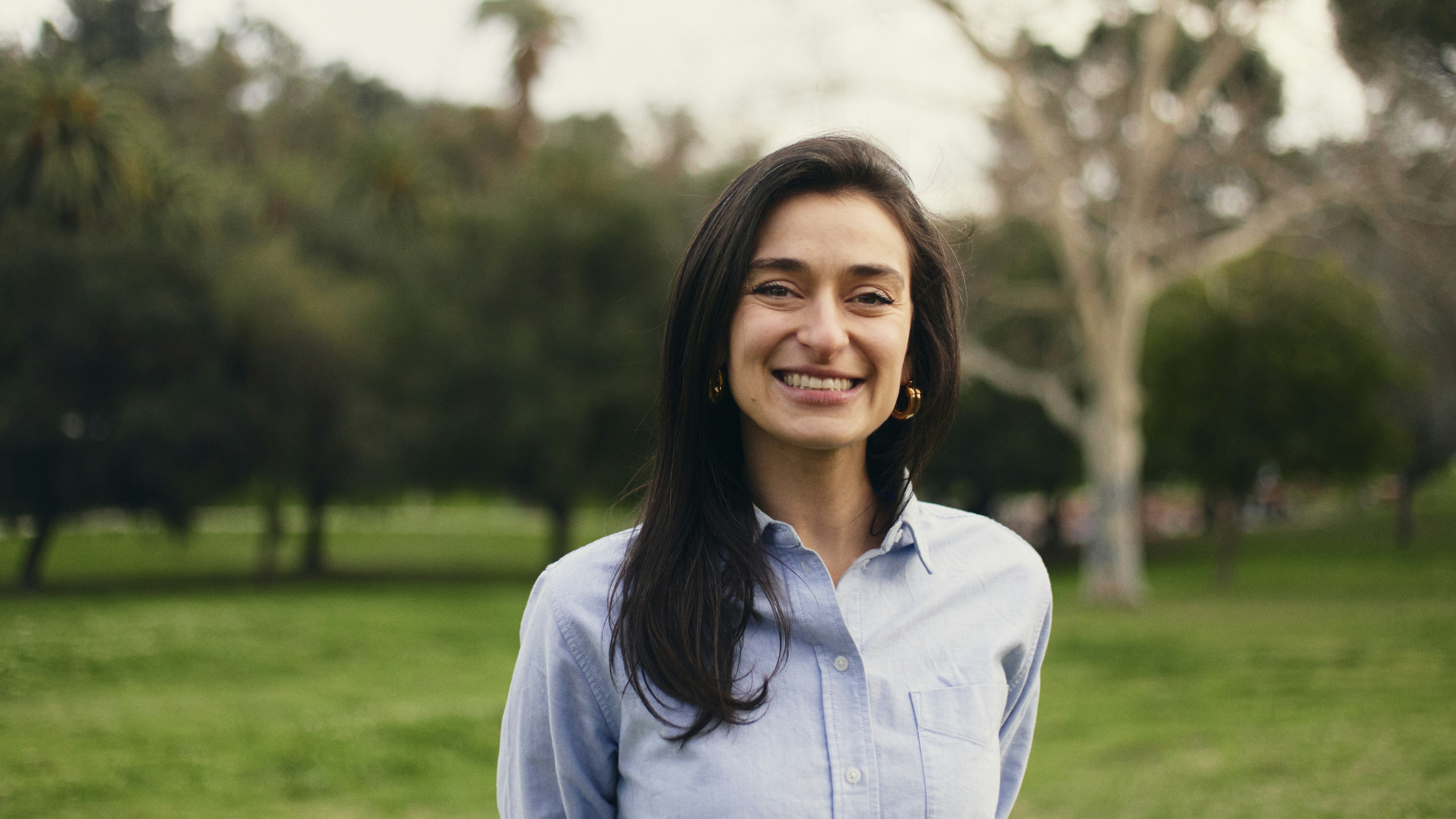 Smiling dark-haired woman wearing light blue button-down shirt stands in a green field.