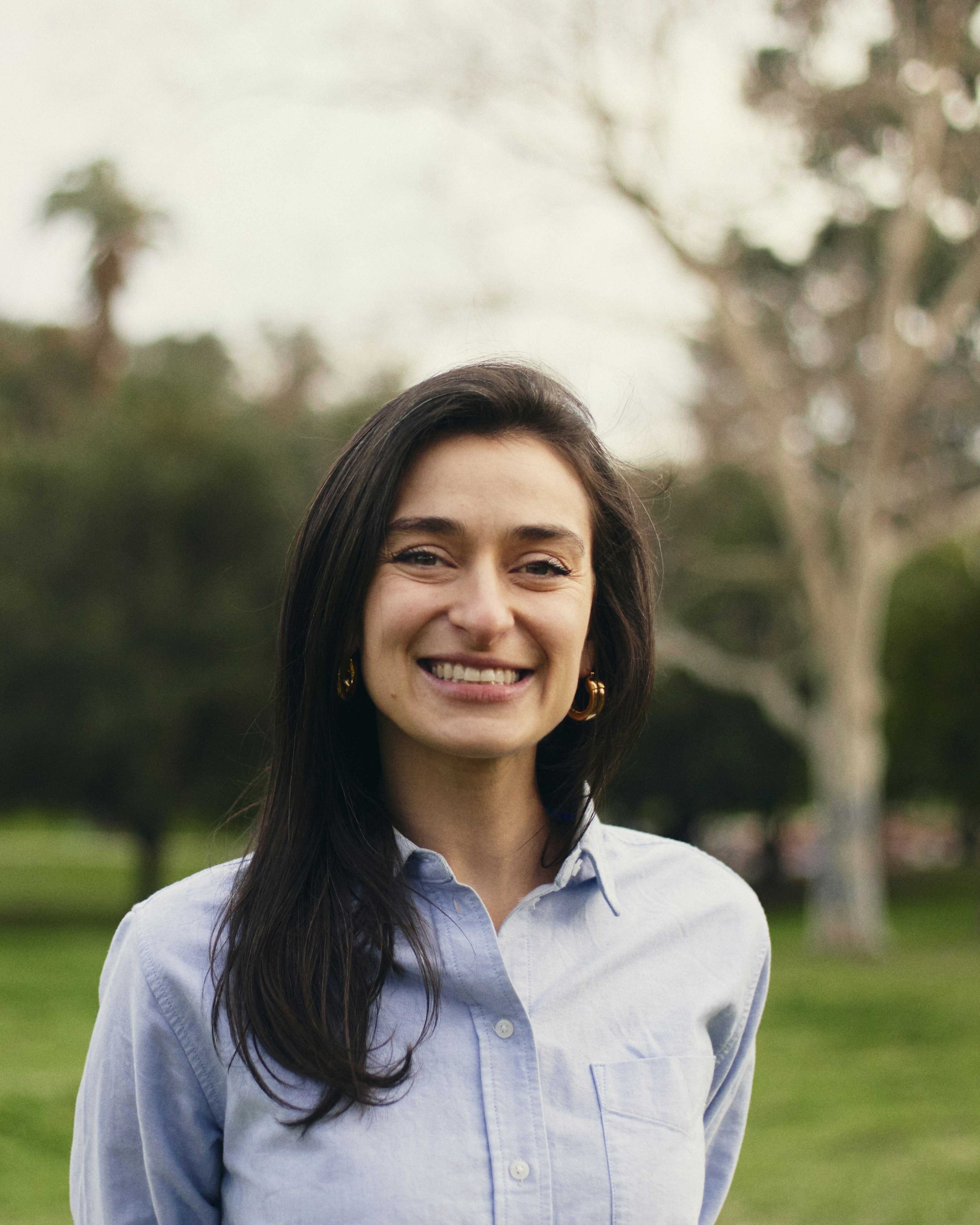 Smiling dark-haired woman wearing light blue button-down shirt stands in a green field.