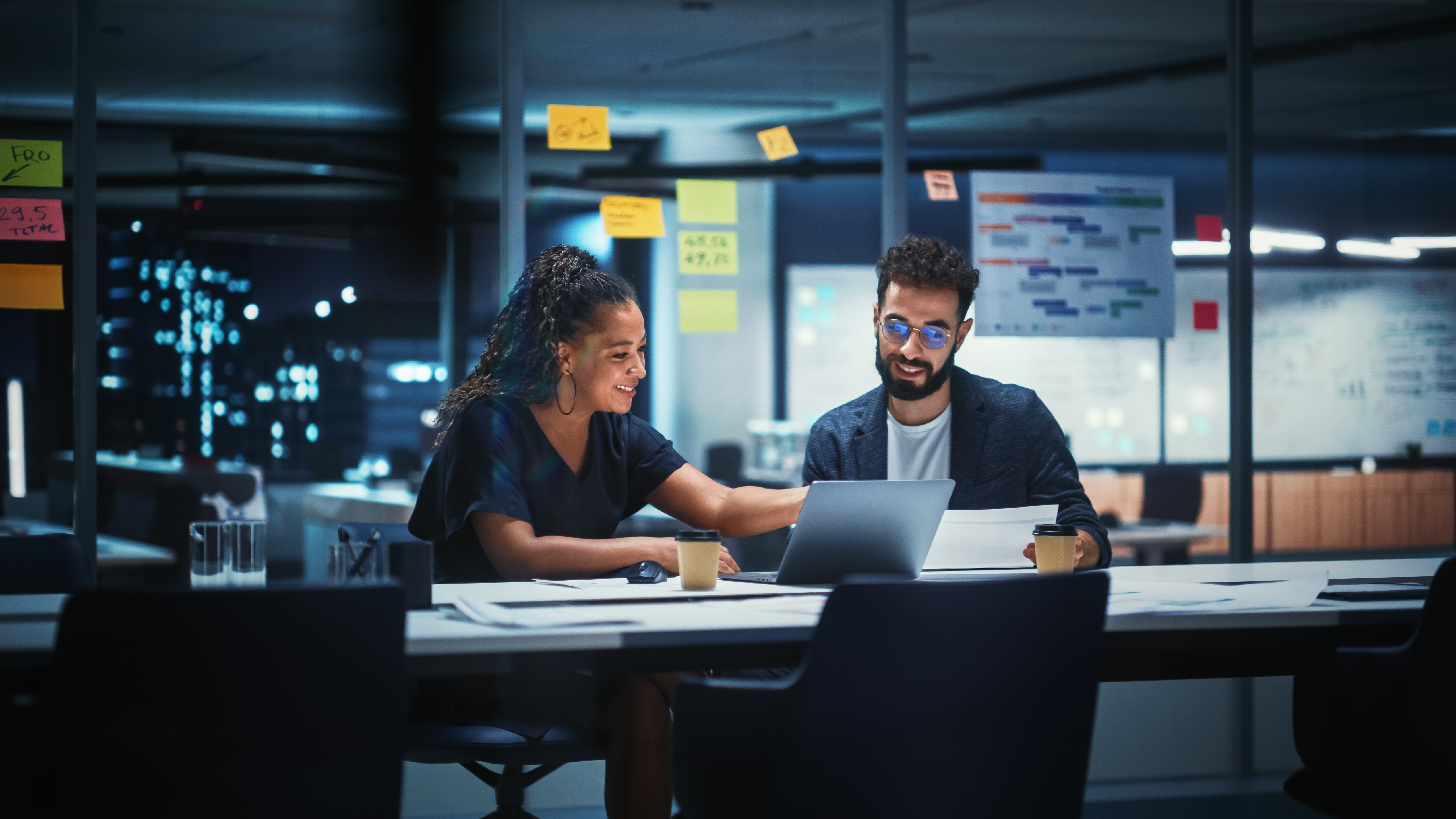 Two colleagues are discussing something, the woman shows something on a laptop. The man is holding a paper