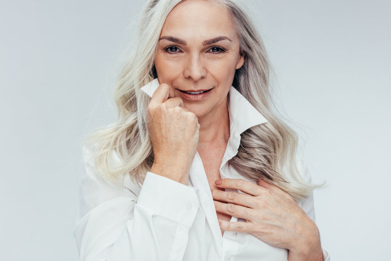 Older woman with long gray hair smiles at the camera in a studio shoot with her collard shirt popped up and holding one side. Her youthful beauty even at her age emulates the potential results of a facelift in Frisco, Plano, Allen, and McKinney
