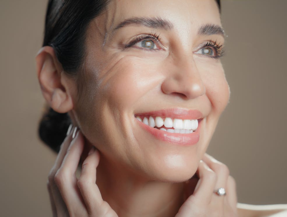 Woman in her 50's with bronw hair smiles wide looking off with her hands underneath her jawline. Her tight skin and defined jaw are to emulate the potential results of a facelift in McKinney with Dr. Chung
