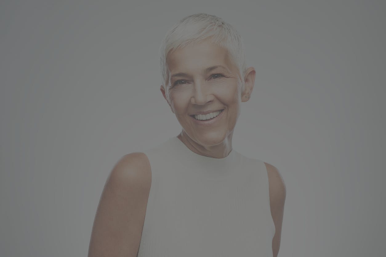 Older woman with long gray hair smiles at the camera in a studio shoot. Her youthful beauty even at her age emulates the potential results of a facelift in Frisco, Plano, Allen, and McKinney
