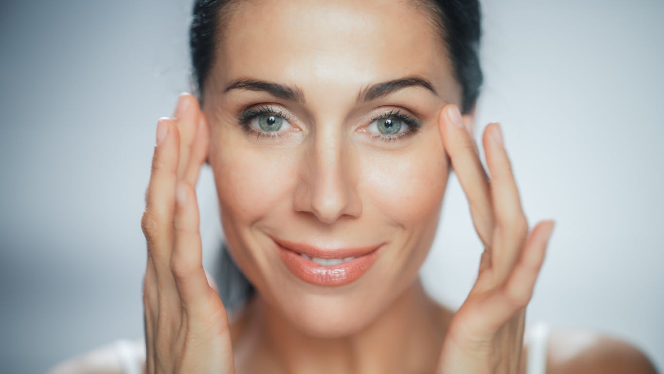 Woman in late 40's with dark brown hair holds hands up to temples. Her youthful appearance is to emulate the possible results of a blepharoplasty in McKinney