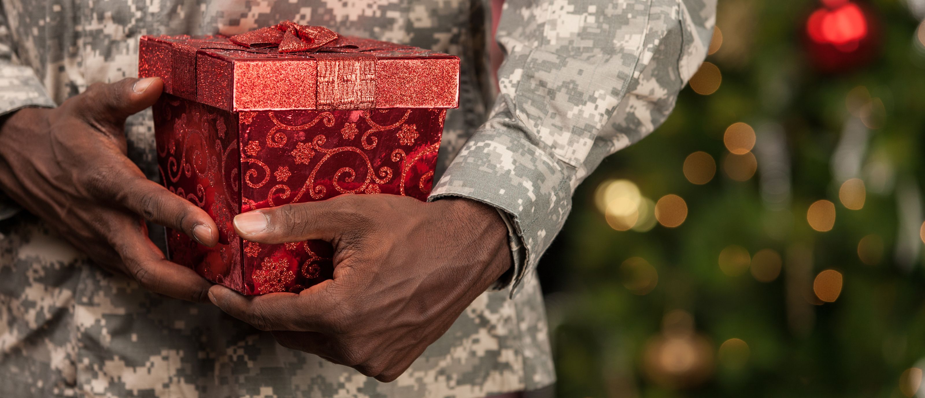 Man in Military uniform holding red present in front of Christmas Tree