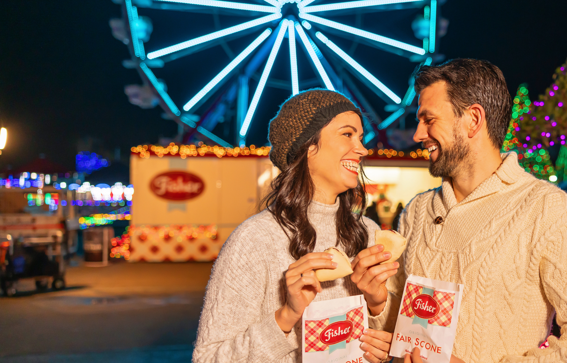 A happy couple in white sweaters eats scones in front of a glowing ferris wheel at Holiday Magic.
