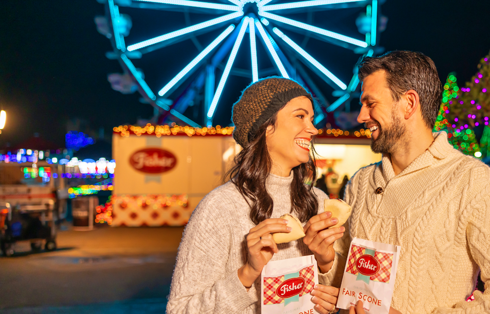 A happy couple in white sweaters eats scones in front of a glowing ferris wheel at Holiday Magic.
