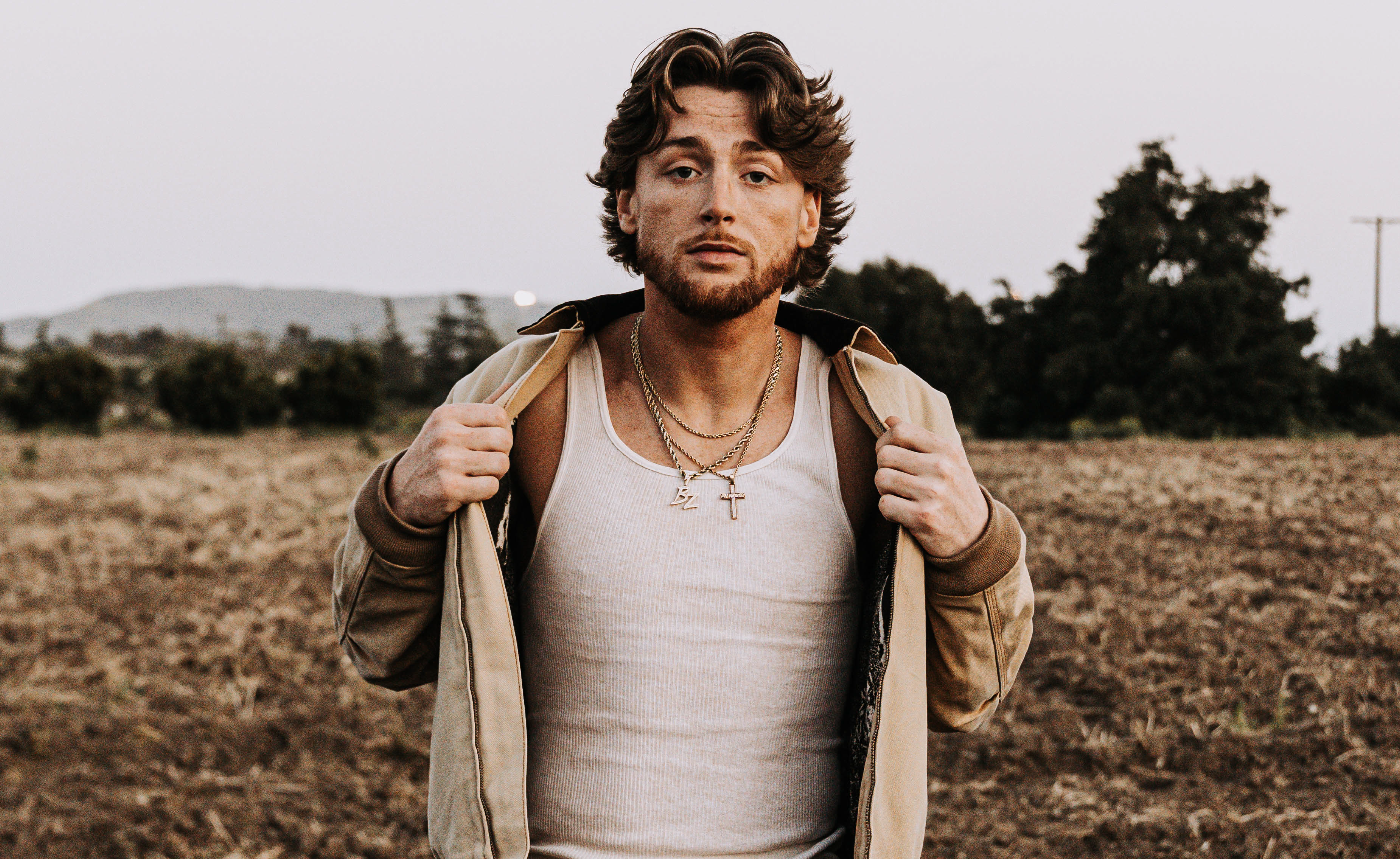 A photo of Bailey Zimmerman wearing a white tank top and tan jacket, standing in a field outdoors.