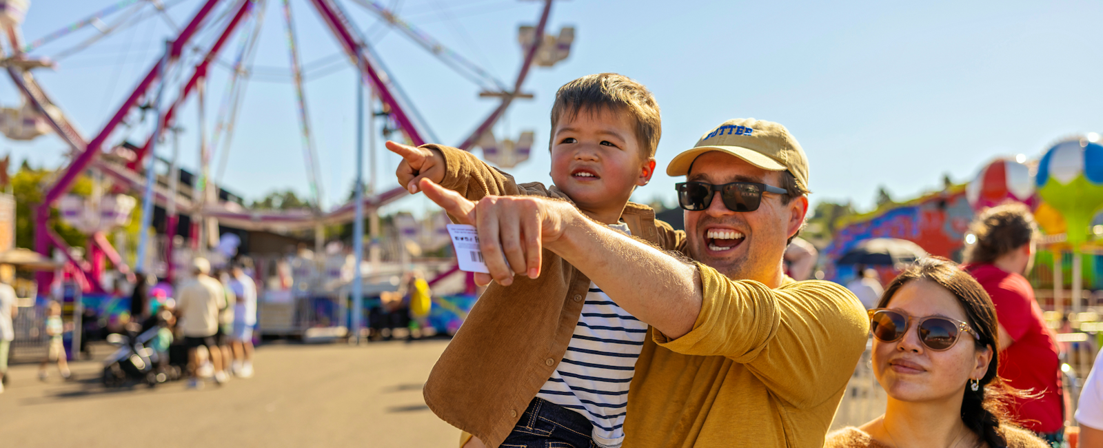 A mom and dad stand in front of a ferris wheel, the dad has their toddler son on his hip and they are pointing out of frame and smiling.