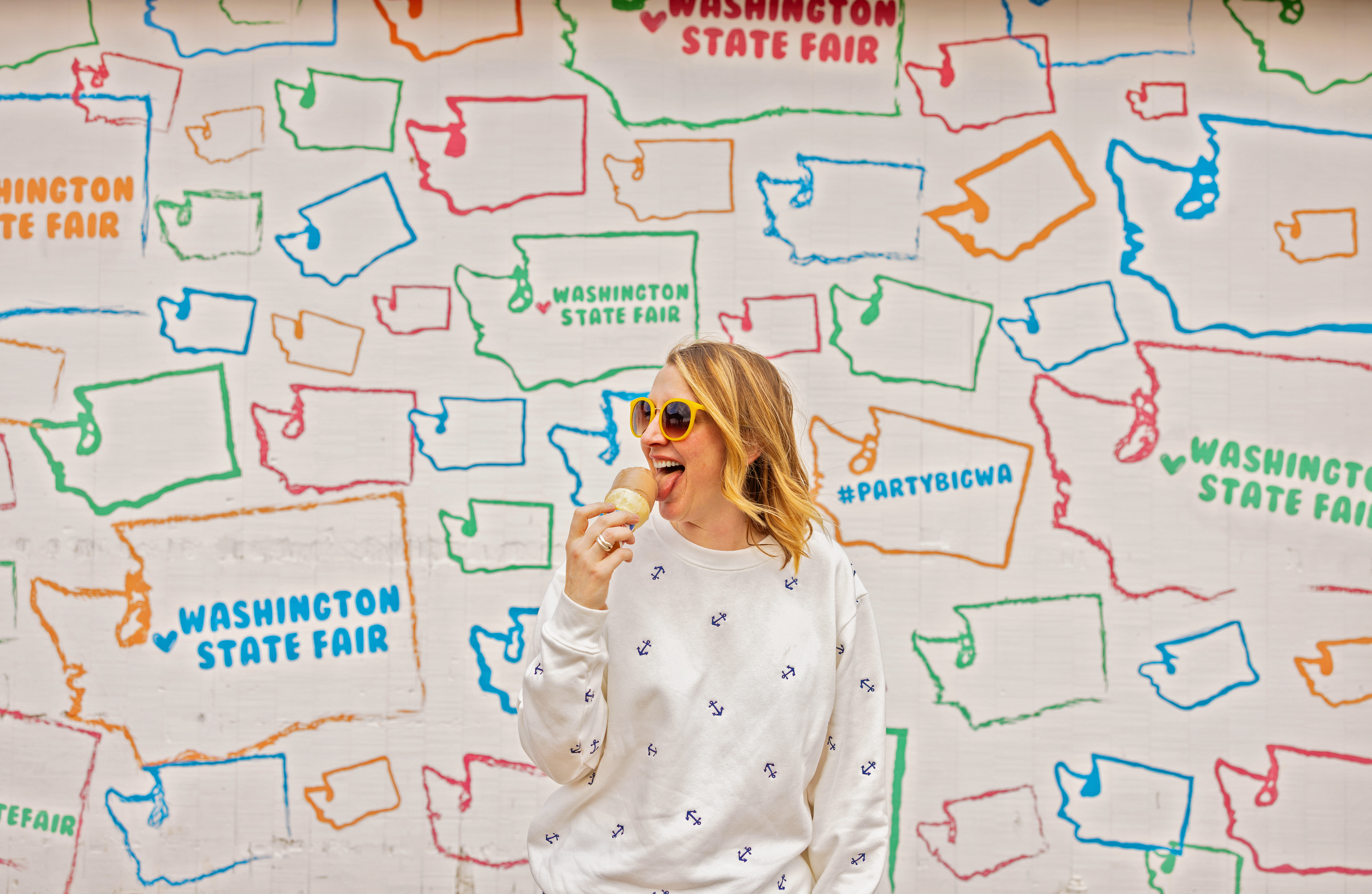 A woman eats an ice cream cone in front of a mural with small, colorful outlines of Washington state.