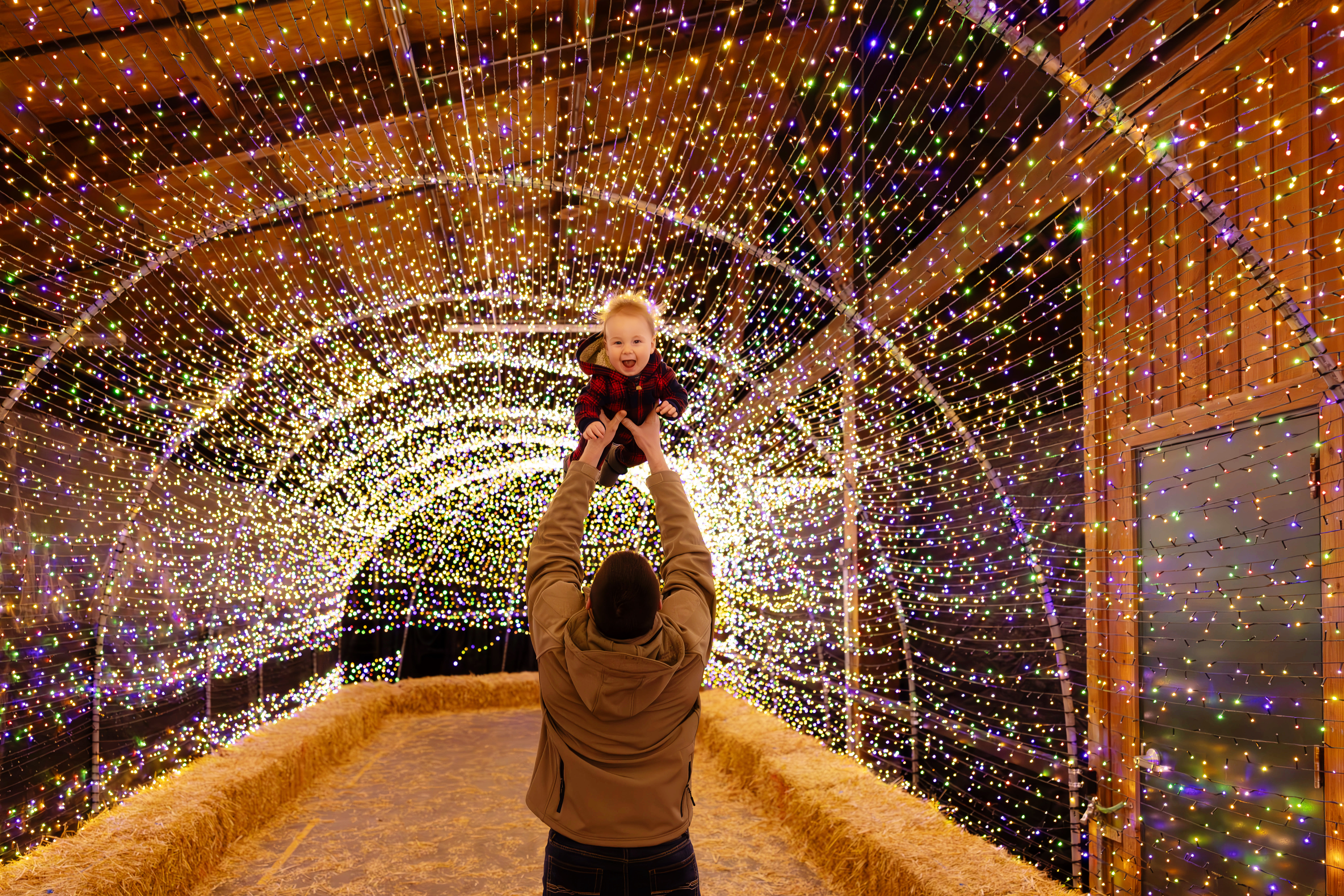 A man holds up his smiling baby in the twinkle light tunnel.