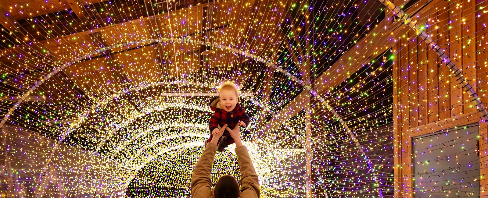 A man holds up his smiling baby in the twinkle light tunnel.