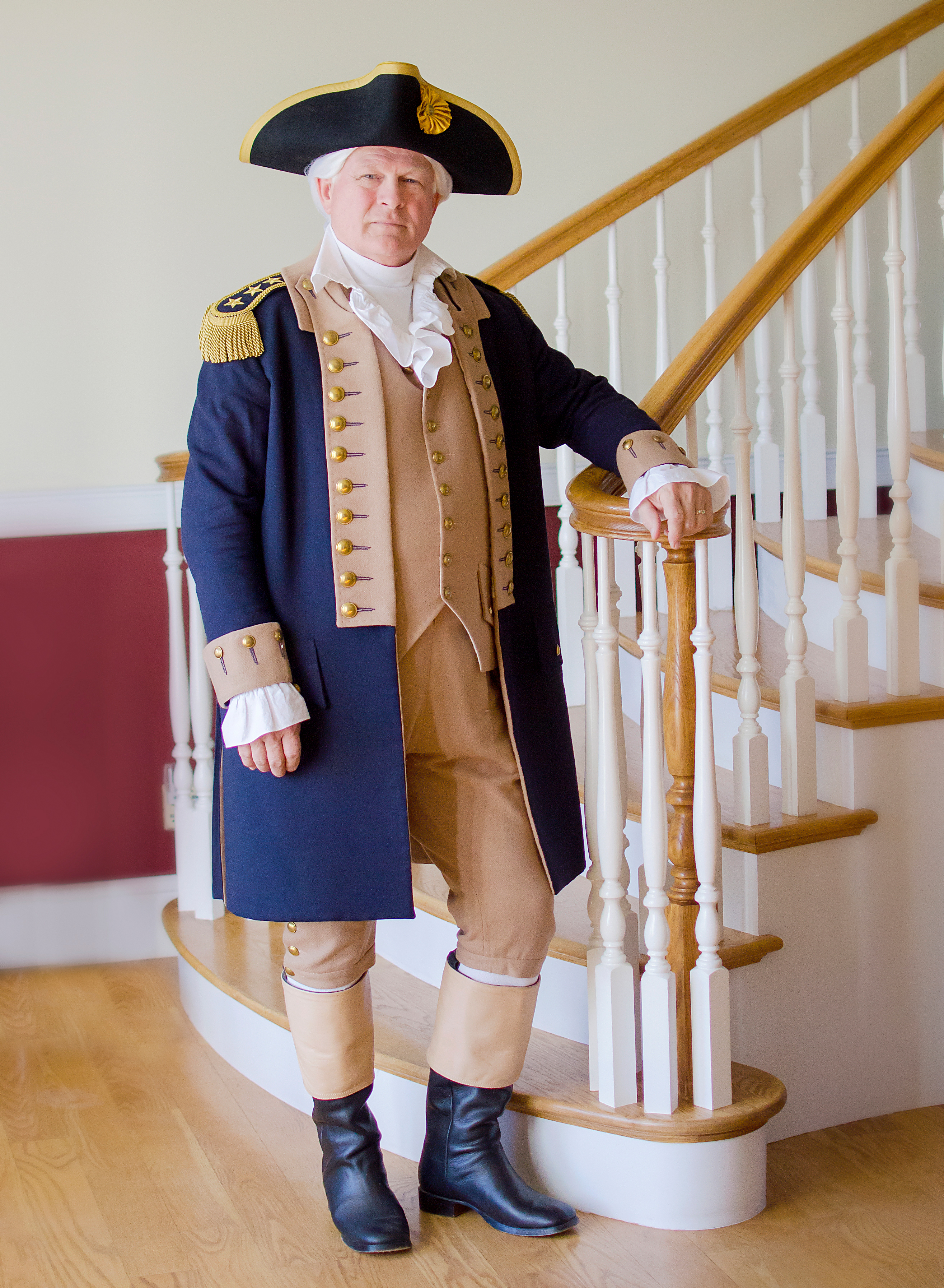 A man dressed as George Washington in a tri-corn hat and tan and blue uniform stands at the bottom of a tall staircase.
