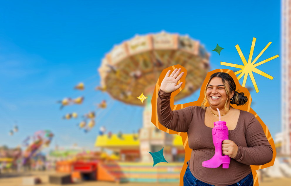 A woman smiling and waving in a brown top and jeans, holding a pink cowboy boot slurpy cup and standing in front of the Wave Swinger ride on a cloudless sunny day.