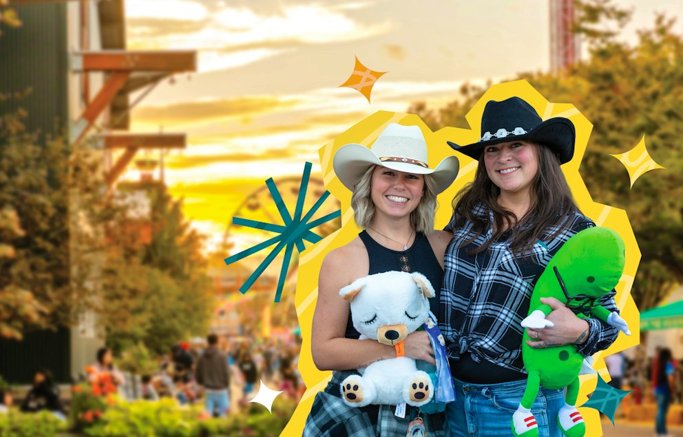 Two friends in black and white cowboy hats hold plushy toy prizes and smile at the camera with the sun setting over the midway in the background.