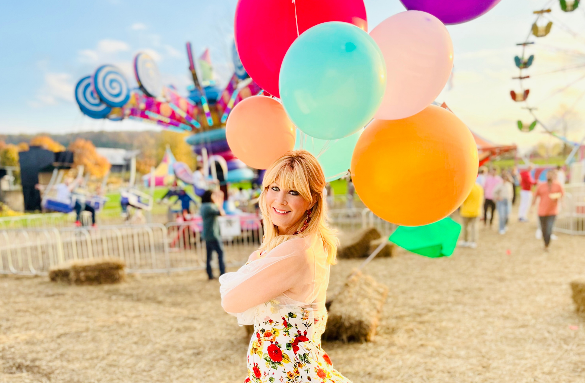 Sandra Lee, a blonde woman smiles at the camera at a fair in front of some carnival rides, holding a bunch of colorful balloons.