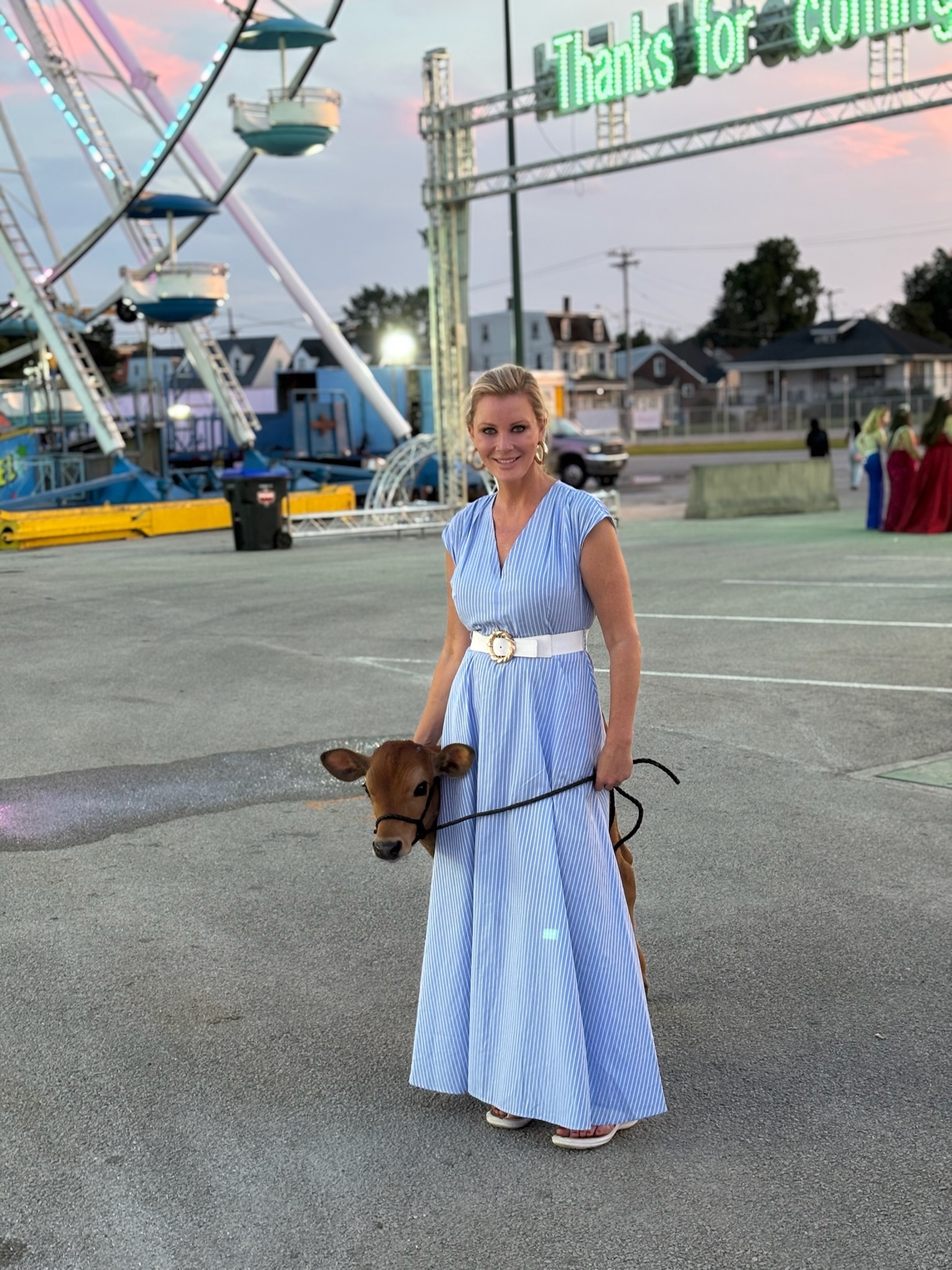 Sandra Lee, a blond woman in a blue dress, stands smiling at the camera at a Fairground with a calf.