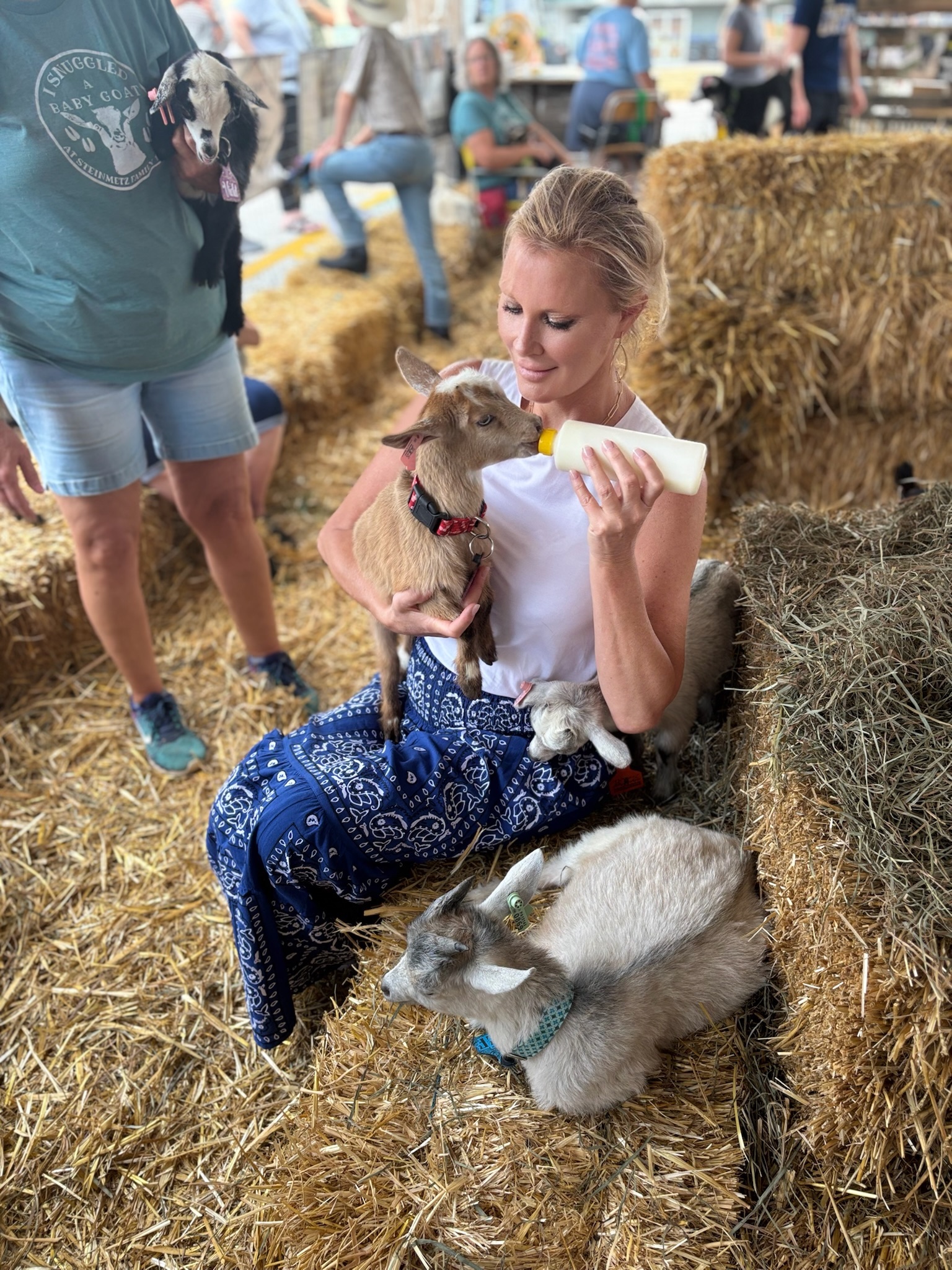 Sandra Lee, a blond woman, sits on a hay barrel holding a baby goat and feeding it from a bottle.