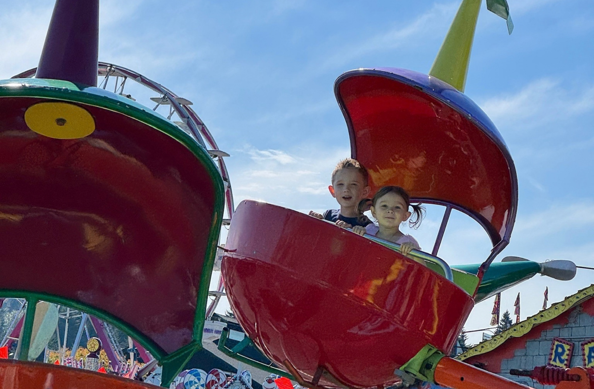 Two children smiing in front of a blue sky while riding Hop-A-Copter, a child's ride with red helicopter-shaped cars that flies them in the air.