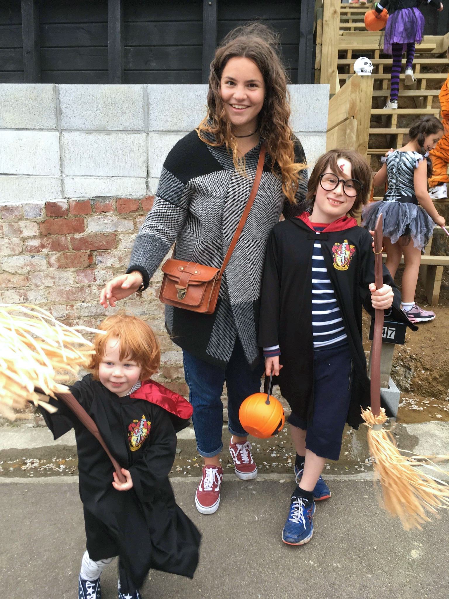 Una ragazza alla pari indossa un abbigliamento casual mentre gioca con due bambini in costume per Halloween, all'aperto in un contesto familiare.