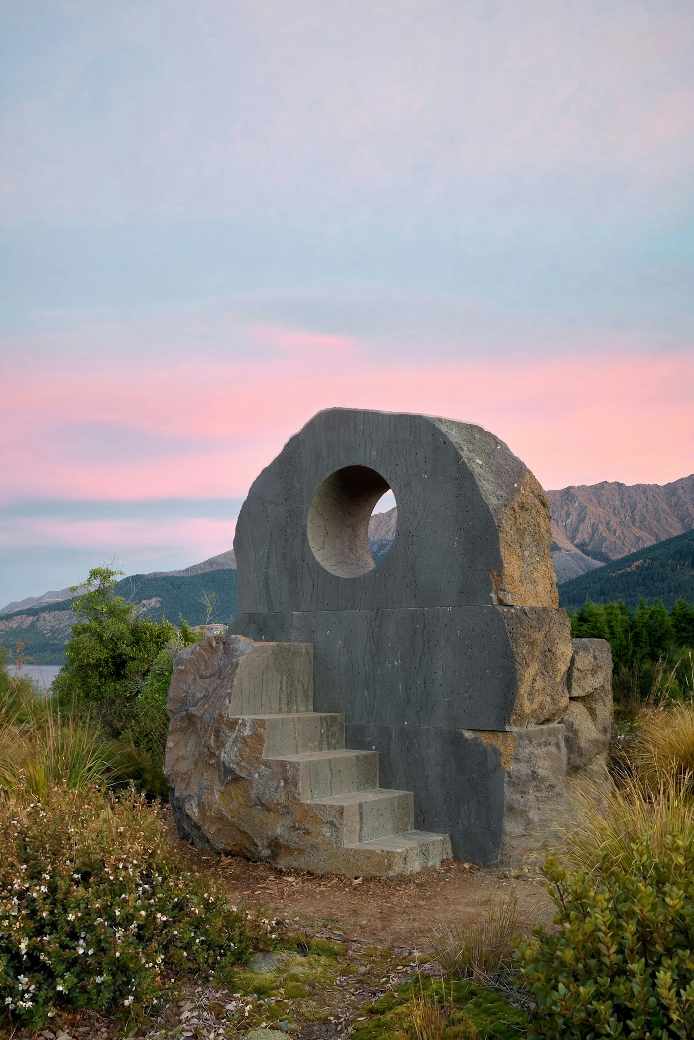 Interlude at dusk: the carved bluestone portal and cast steps on the shore of Lake Whakatipu, the Remarkables and a pink evening sky behind.