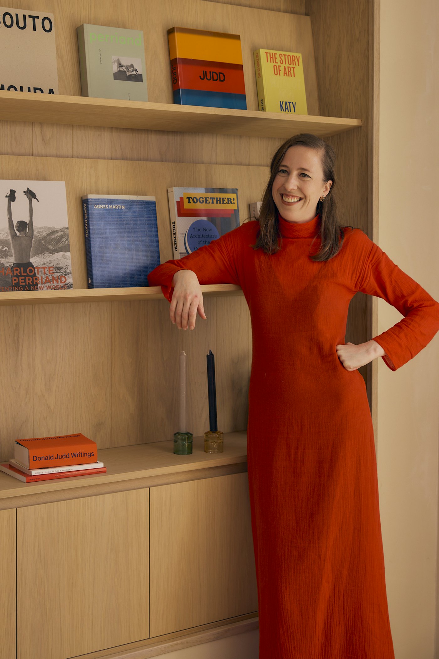 Raphaela Rose, smiling, in a long red dress with one arm resting on a wooden bookshelf of art and architecture books.