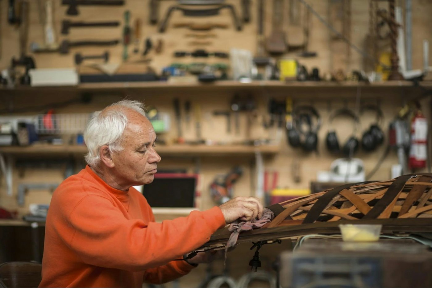Carin Wilson, a white-haired man in an orange jumper, polishing a curved timber frame in his workshop, with tools hung on the wall behind him.