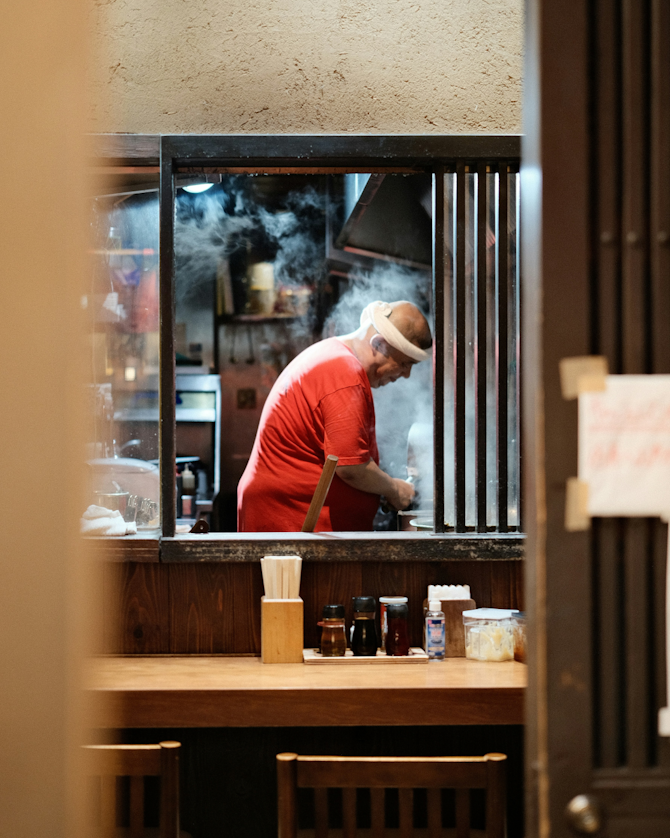 A person in a red shirt works in a steamy kitchen, viewed through a window.