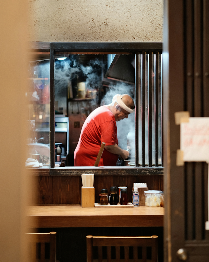 A person in a red shirt works in a steamy kitchen, viewed through a window.