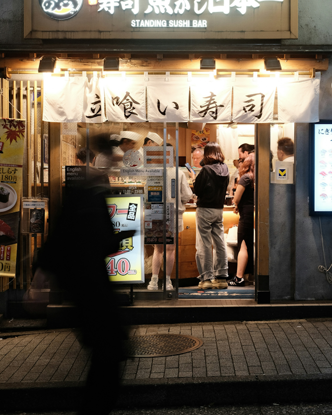 People standing and eating inside a sushi bar, viewed from the street. A blurred pedestrian walks by in the foreground.